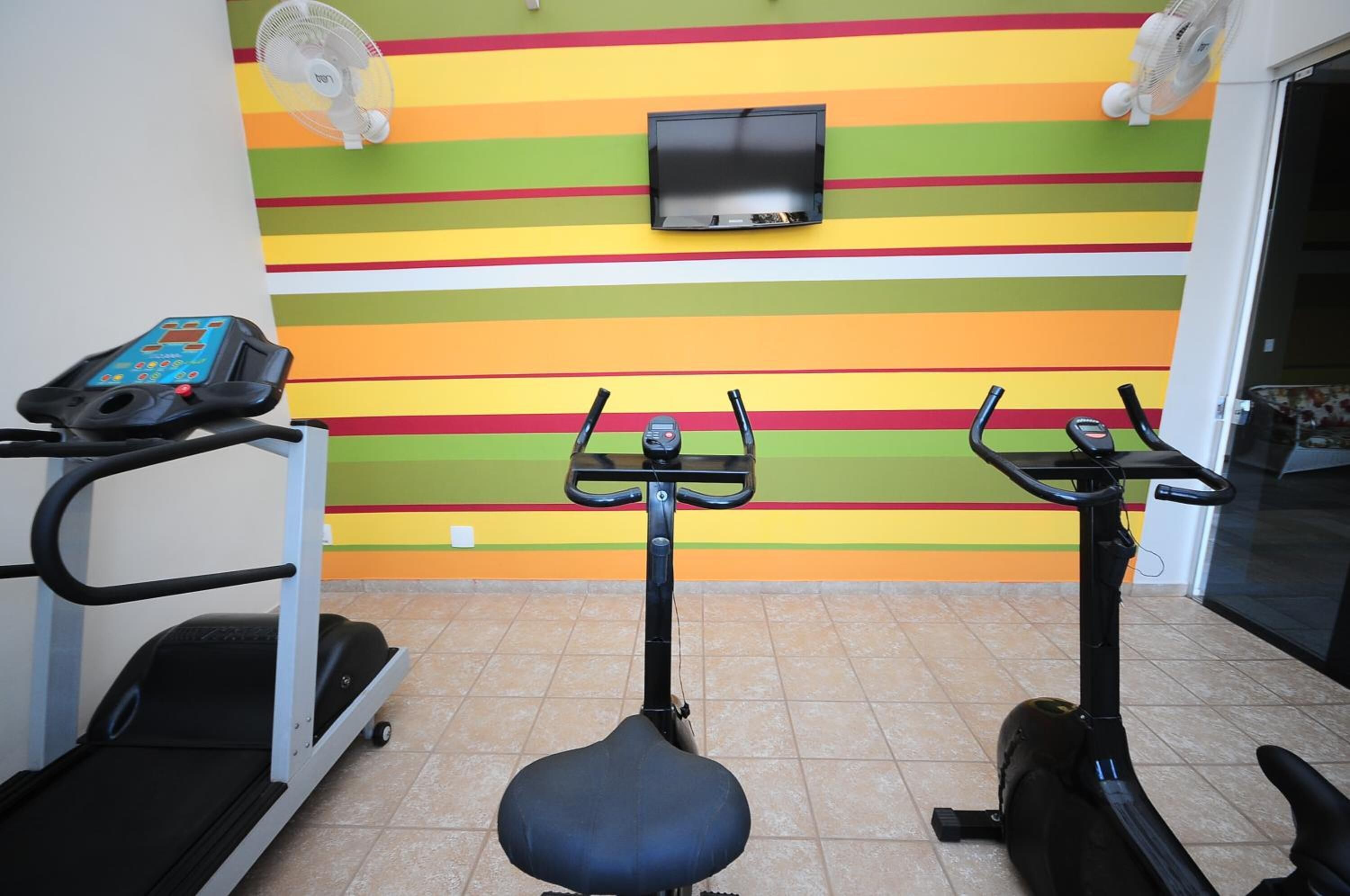 Gimnasio interior con equipo de ejercicio, pared con rayas de colores y un televisor montado.