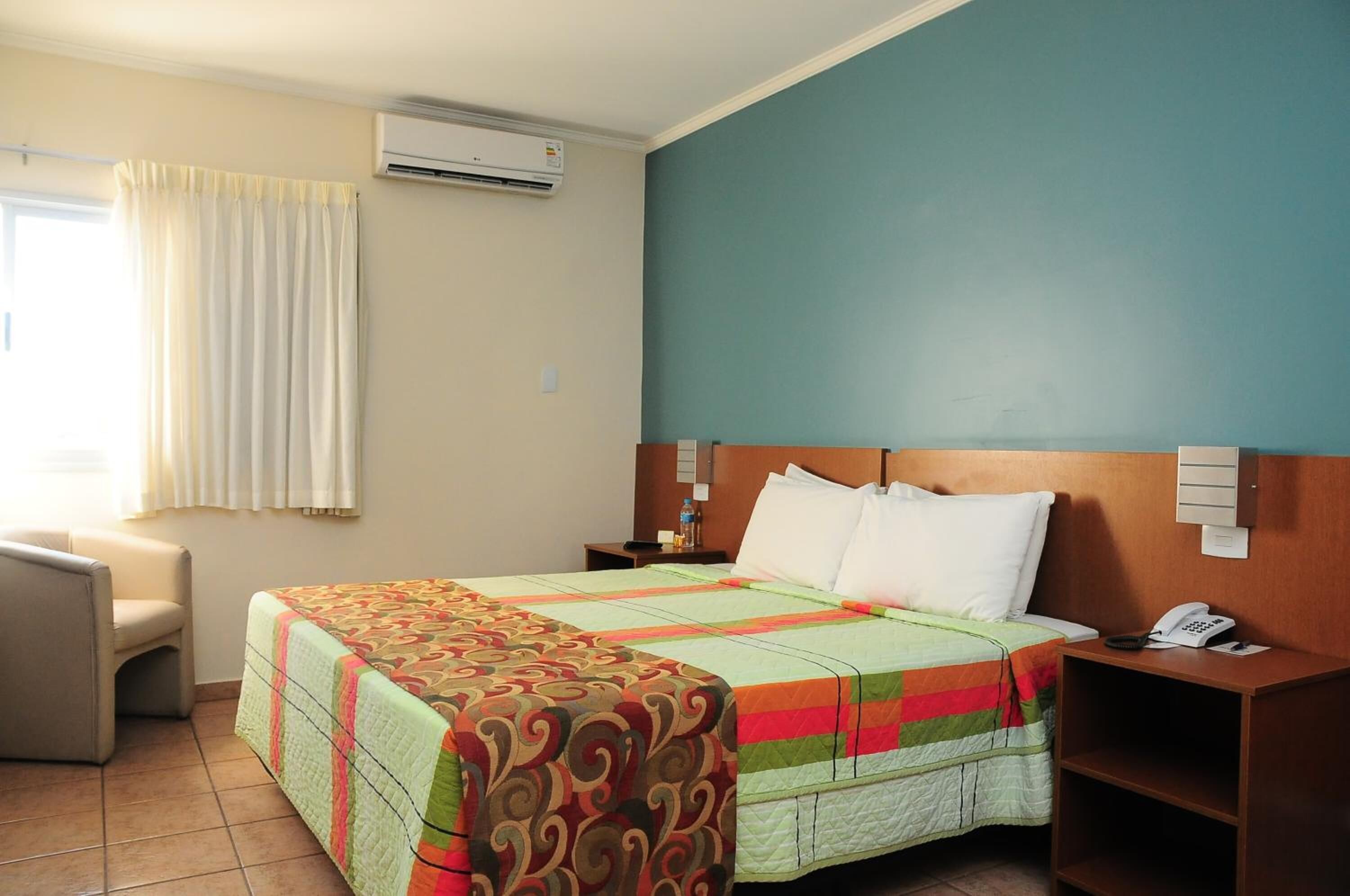 A hotel room with a bed, a chair, a nightstand with a phone, and an air conditioning unit.