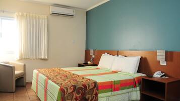 A hotel room with a bed, a chair, a nightstand with a phone, and an air conditioning unit.