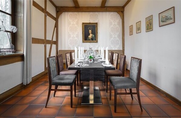 A dining room with a long table set for six, surrounded by chairs, with a framed portrait on the wall and a window with curtains.
