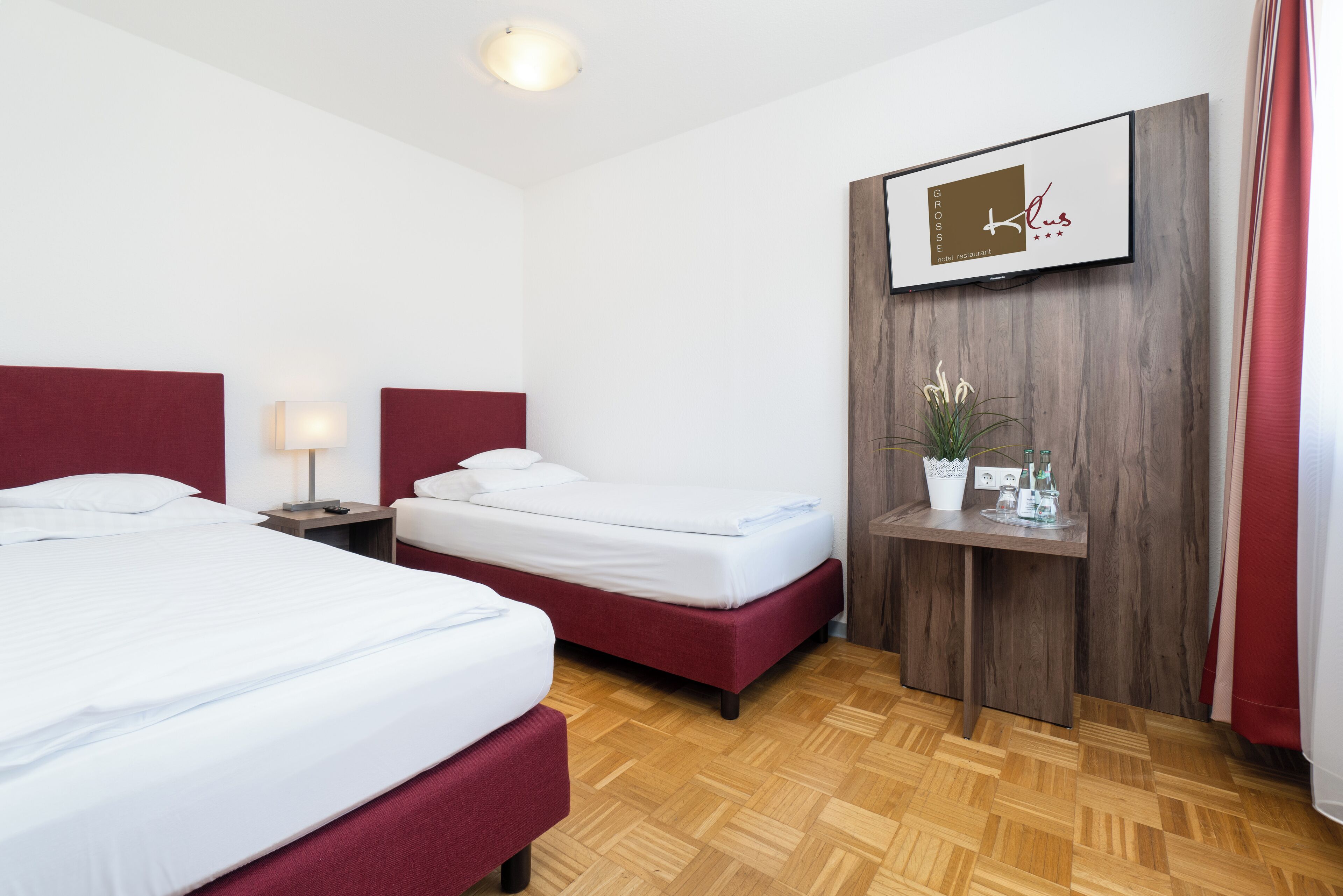 A hotel room with two beds, a wooden headboard, a small table with a plant, and a wall-mounted TV.