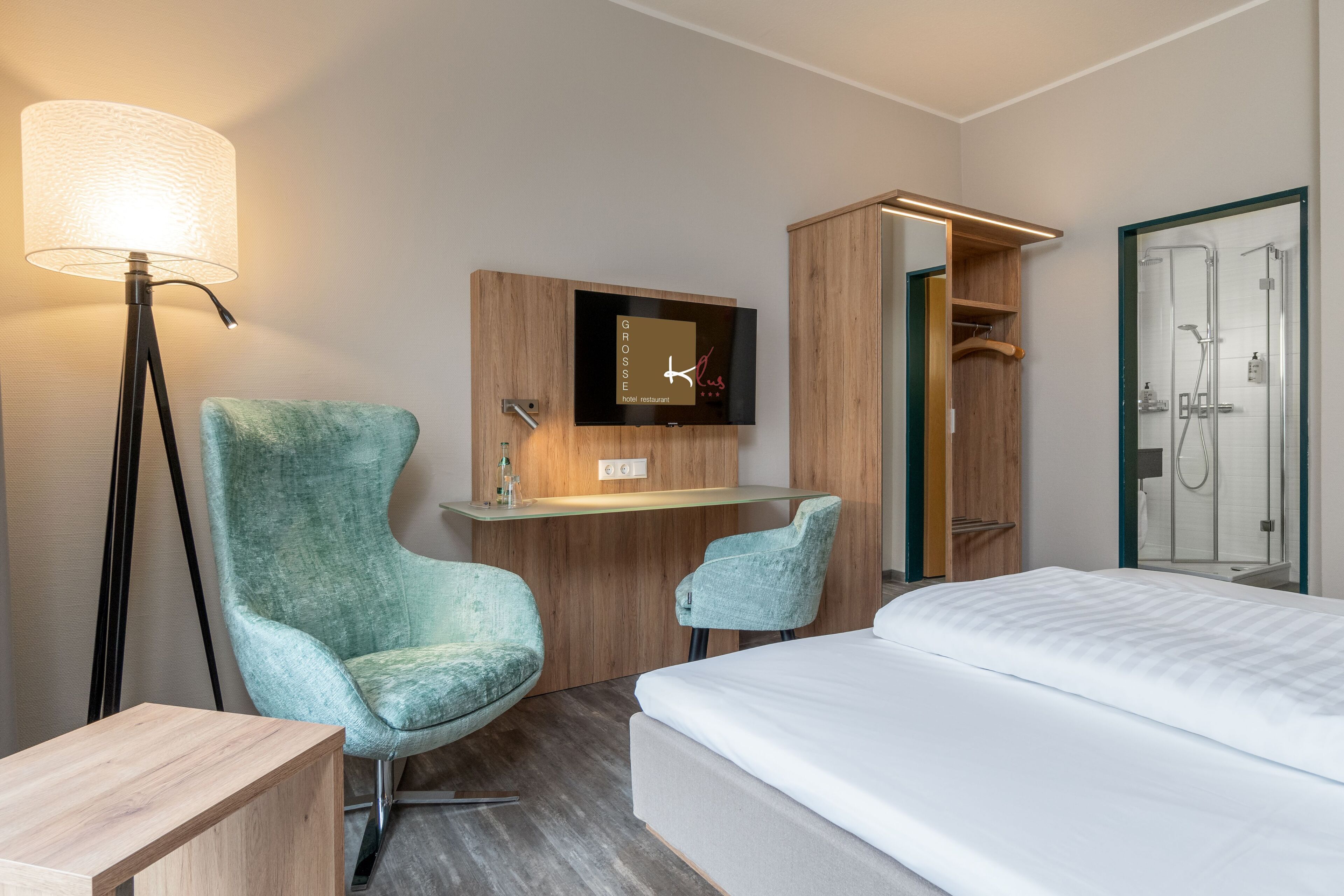 A hotel room with a bed, a desk with a TV, a chair, a lamp, and a shower area.