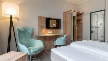 A hotel room with a bed, a desk with a TV, a chair, a lamp, and a shower area.