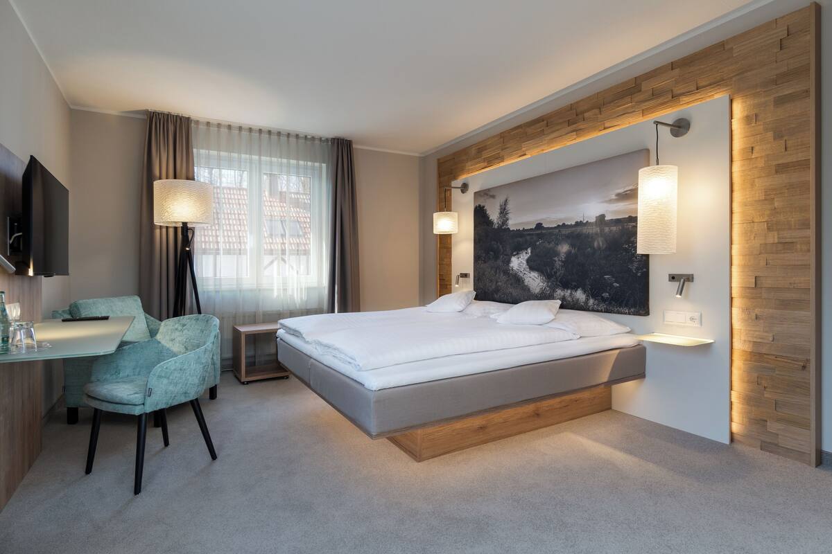 Une chambre d’hôtel moderne, équipée d’un lit, d’un bureau, d’une chaise, d’une télévision et ornée d’un grand tableau représentant un paysage.