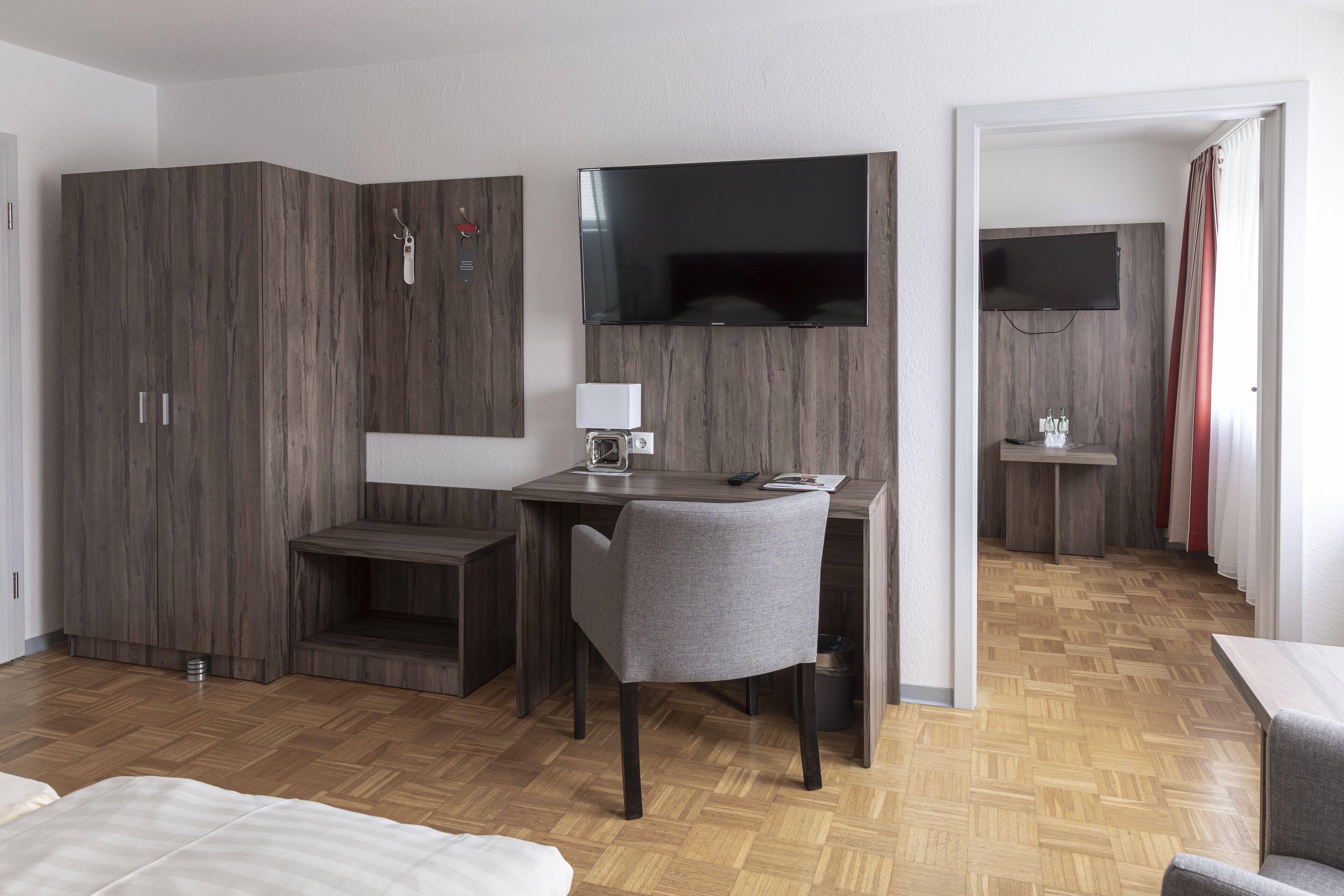 A modern hotel room with a wooden wardrobe, a desk with a chair, a TV mounted on the wall, and a small table with a lamp.