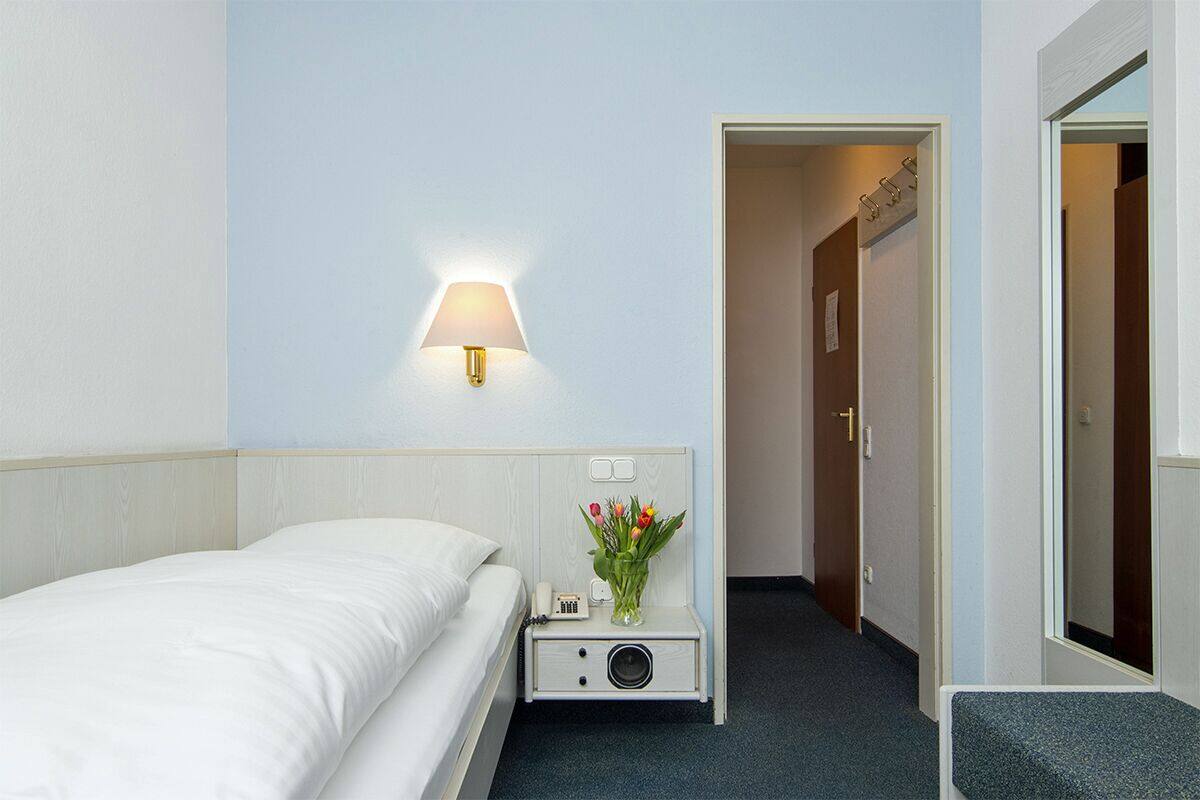 A hotel room with a bed, a bedside table with a lamp, a phone, and a vase with flowers. There is a door leading to another room.