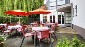 An outdoor patio with red chairs and tables, a red umbrella, and a building with a tiled roof in the background.