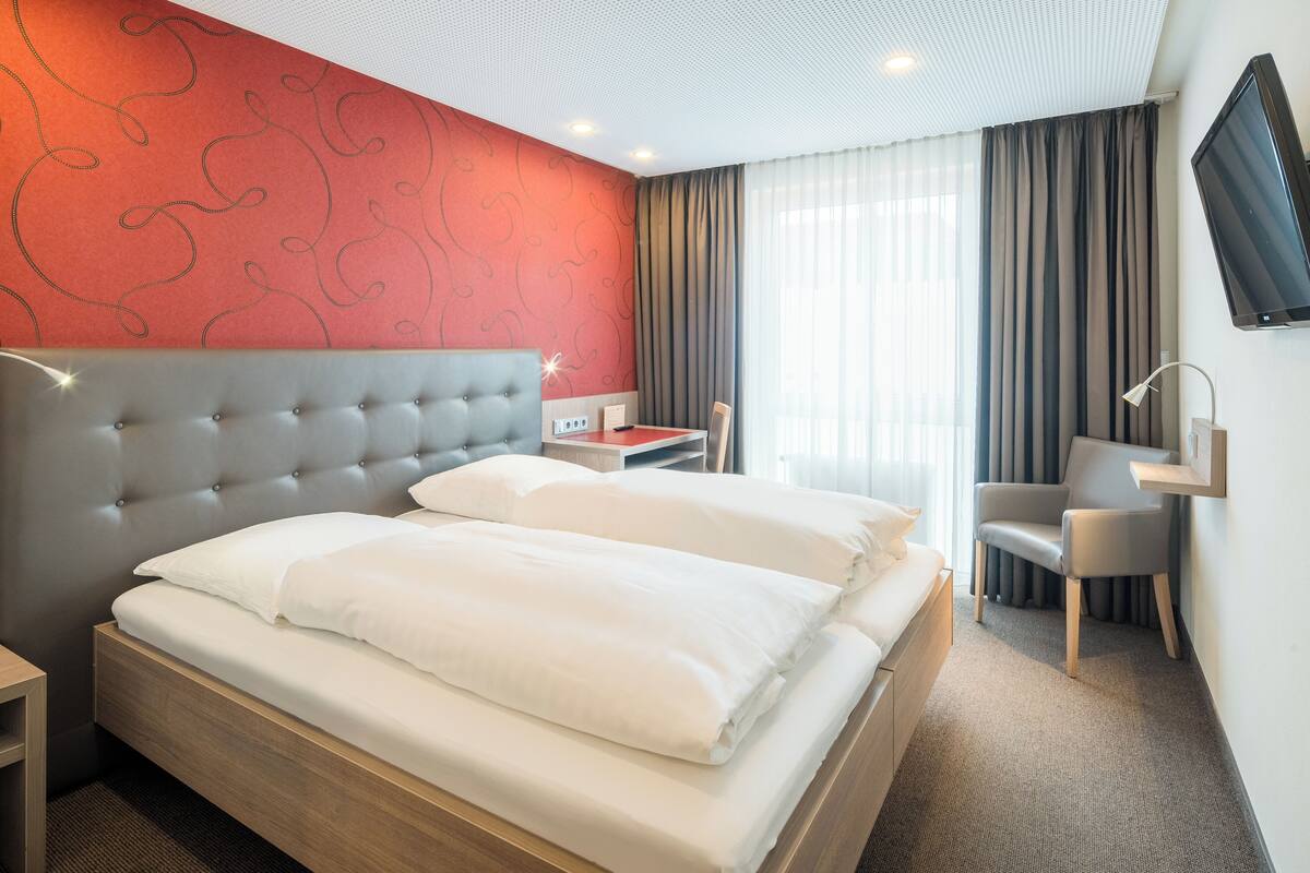 A hotel room with a large bed, a desk, a chair, and a TV on the wall.