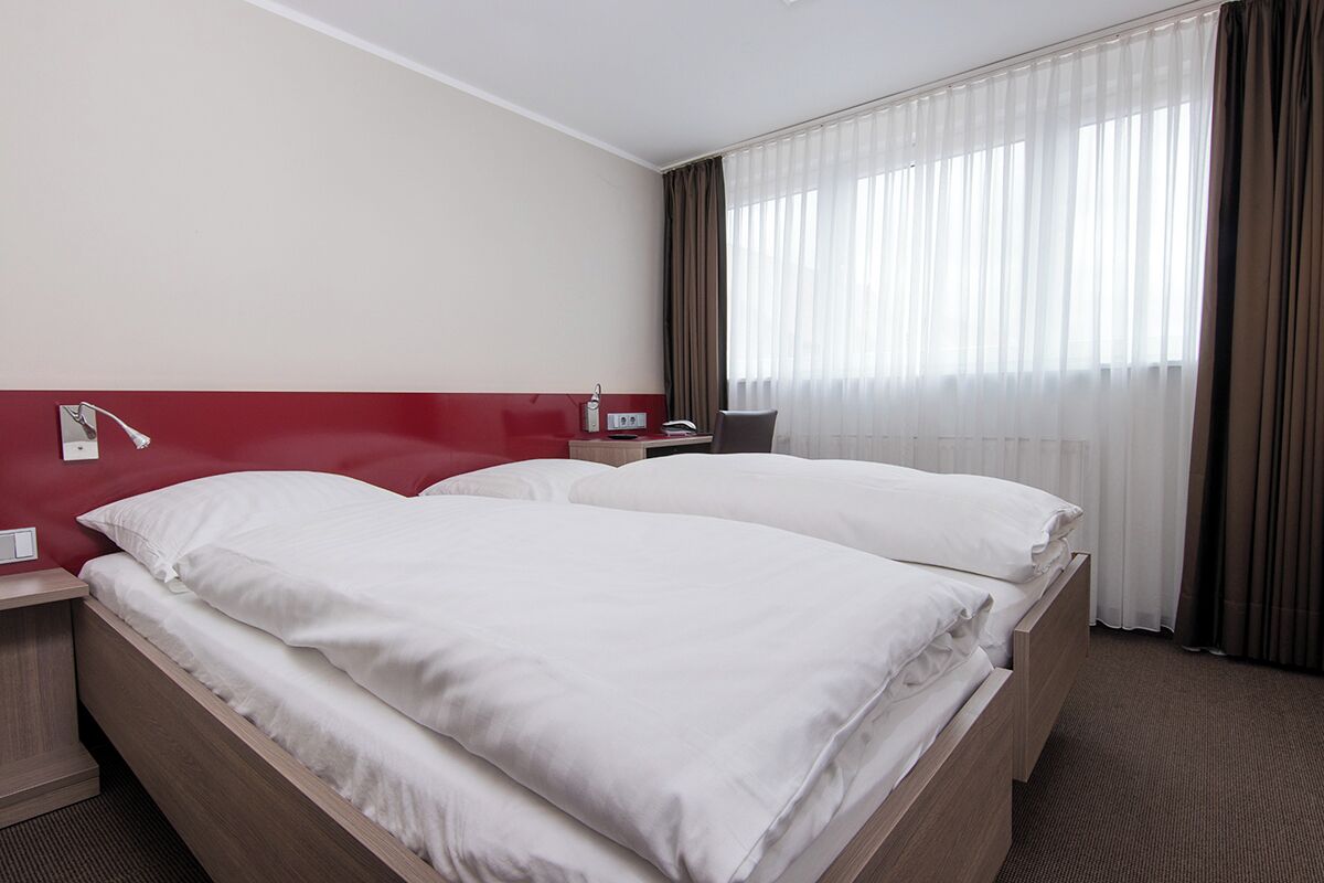 Ein Hotelzimmer mit Doppelbett, Nachttisch, Lampe und einem Fenster mit Vorhängen.