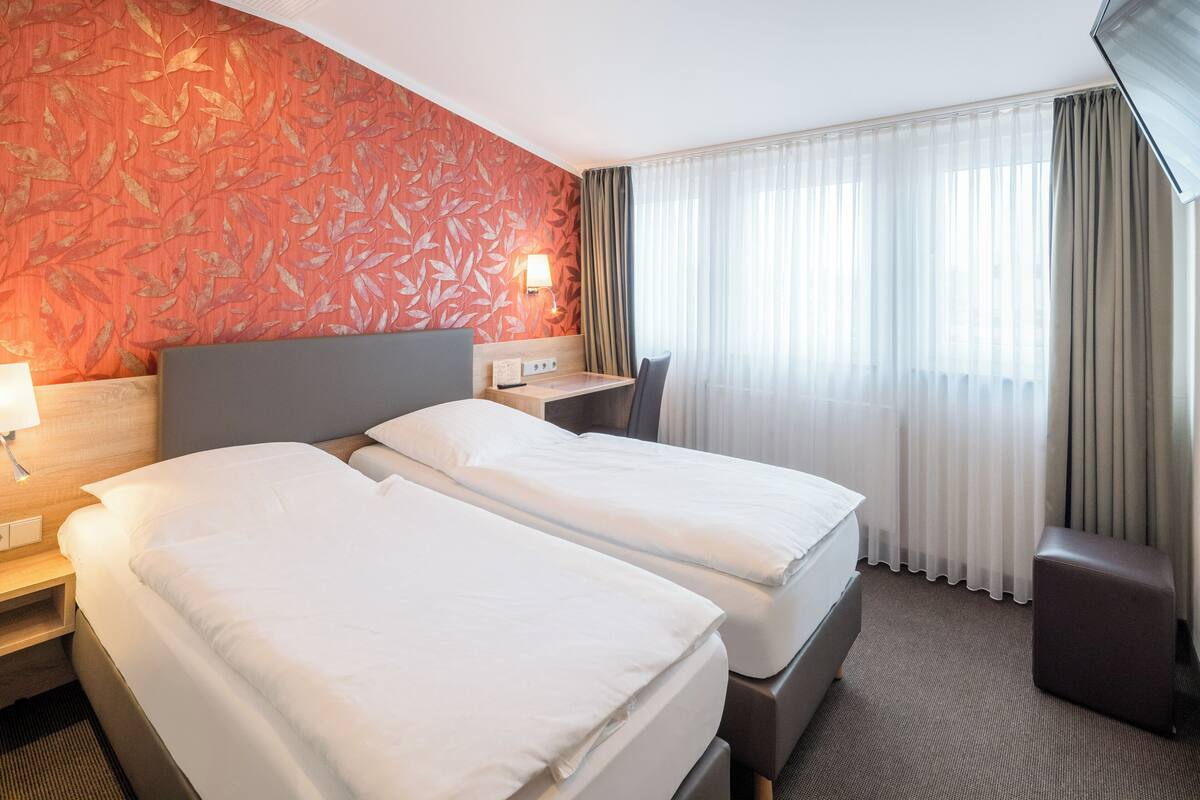 A hotel room with two beds, a desk, a chair, and a large window with curtains.
