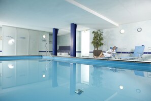 An indoor swimming pool with clear water, a blue and white color scheme, and a reception area with a plant and seating.