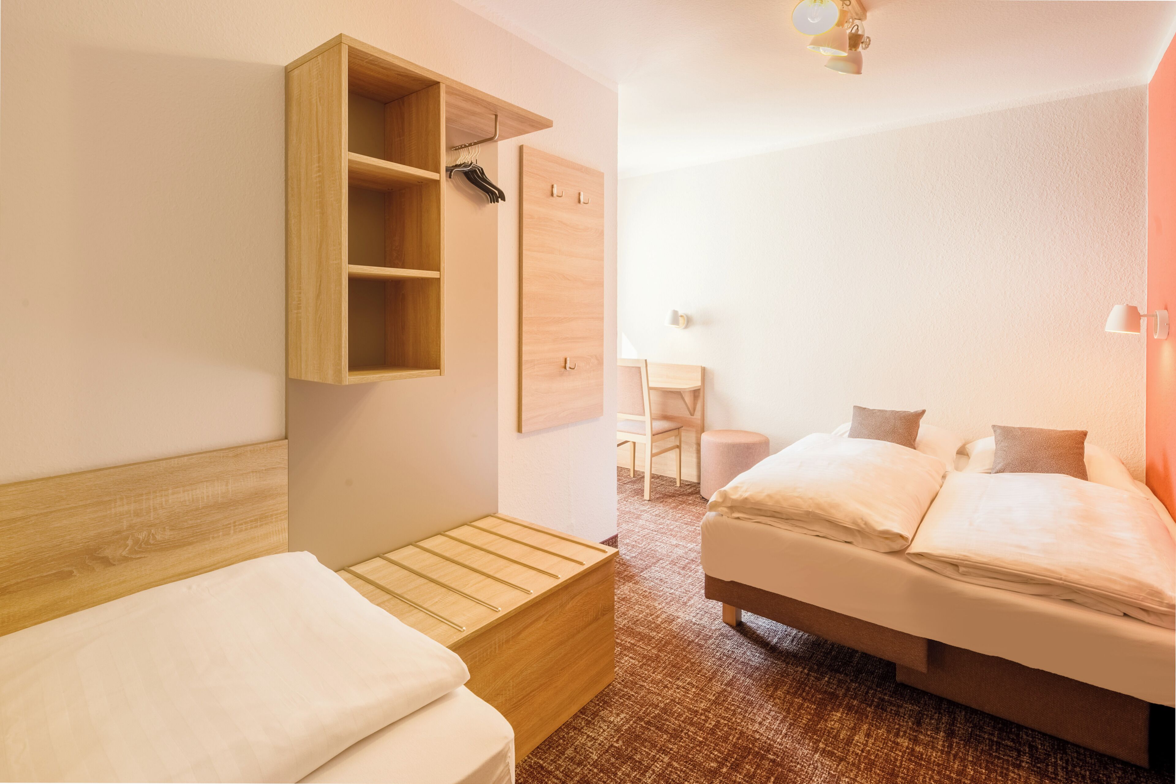 Una habitación de hotel con cabecera de madera, una cama con ropa de cama blanca, un pequeño banco de madera, un escritorio con silla y un estante con perchero.