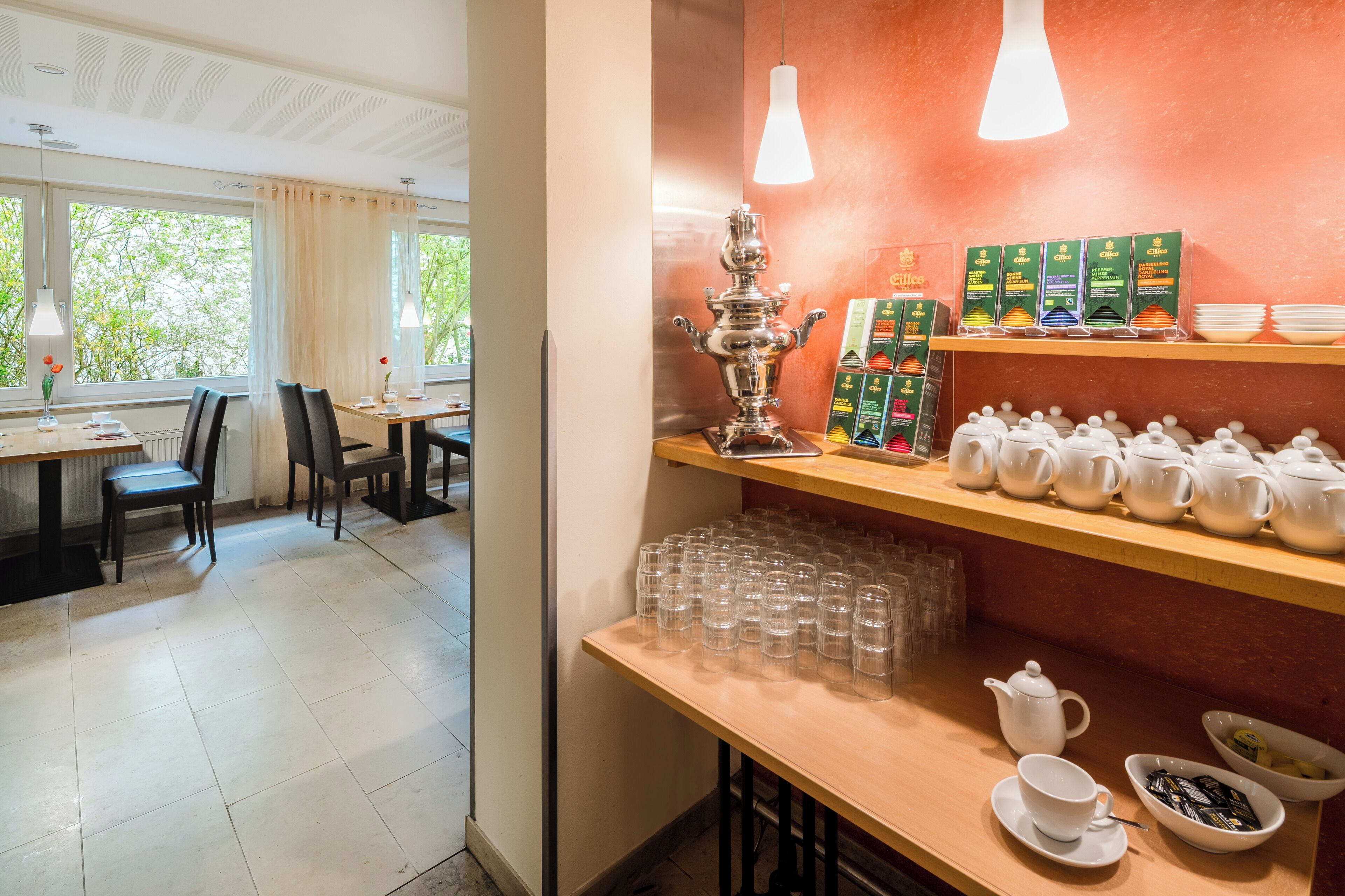 Un coin repas avec une table dressée pour deux personnes, un espace dédié à la préparation du thé avec des théières et des tasses, et une vue sur l’extérieur grâce à de grandes fenêtres.