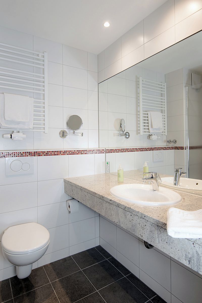 Une salle de bain moderne avec des carreaux blancs, un comptoir en granit, deux lavabos, des toilettes et un grand miroir.