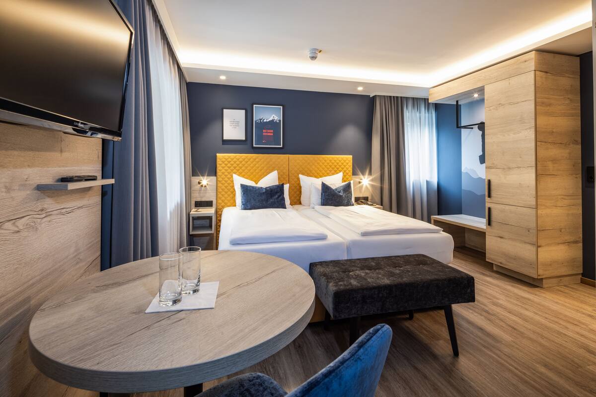 A modern hotel room with a large bed, a round table with two glasses, a TV mounted on the wall, and a wooden cabinet.