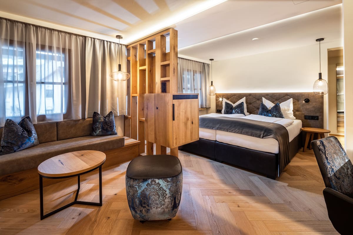 A modern hotel room with a sofa, a round wooden table, a large bed with a headboard, and a wooden wardrobe.