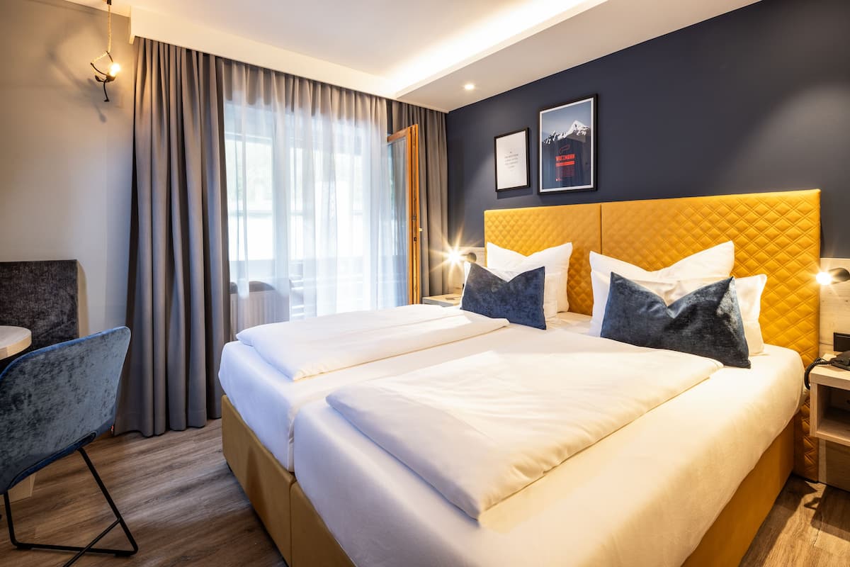 A hotel room with two beds, a desk, a chair, and a large window with curtains.
