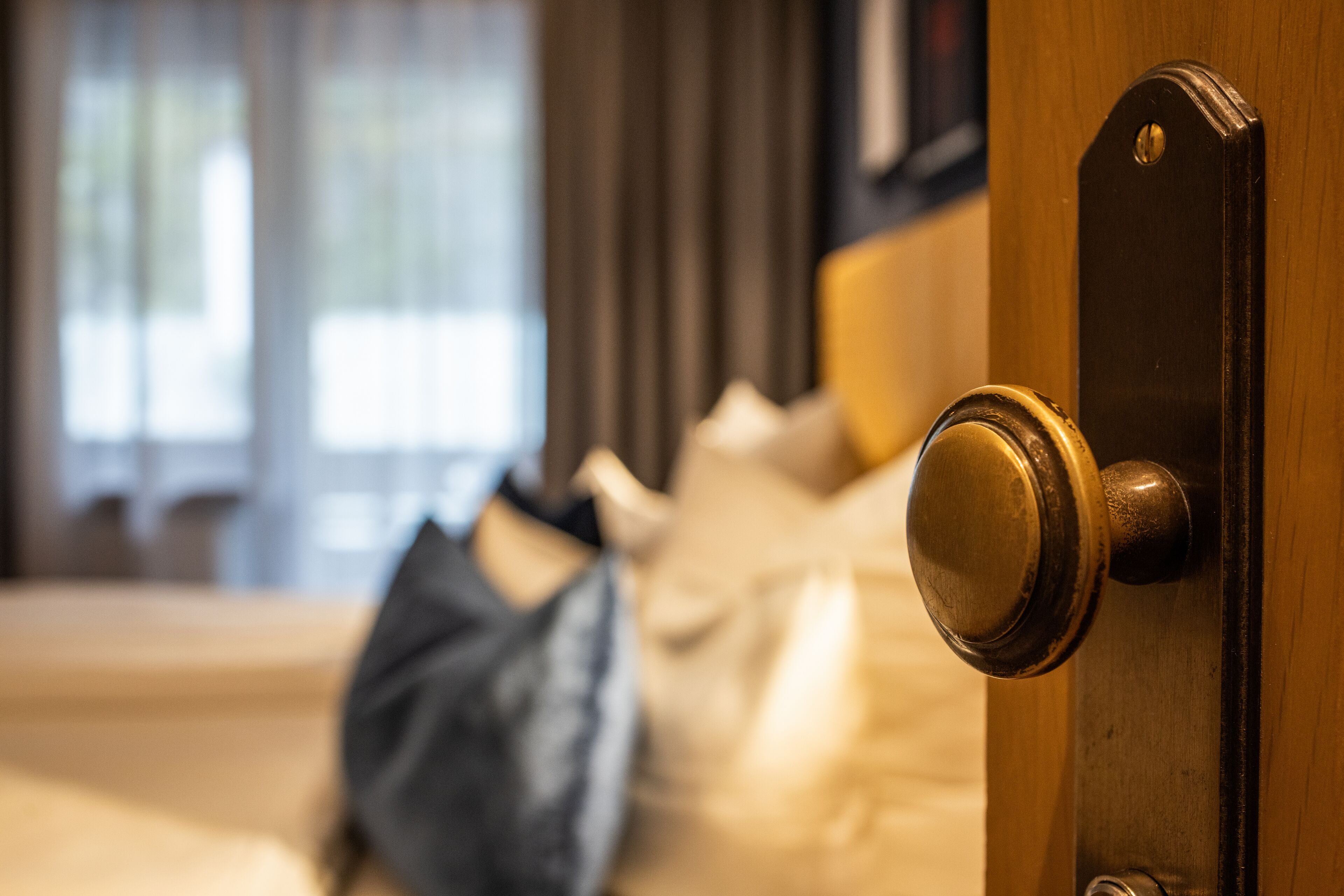 A hotel room with a wooden door, a bed with a white comforter, and a window with curtains.