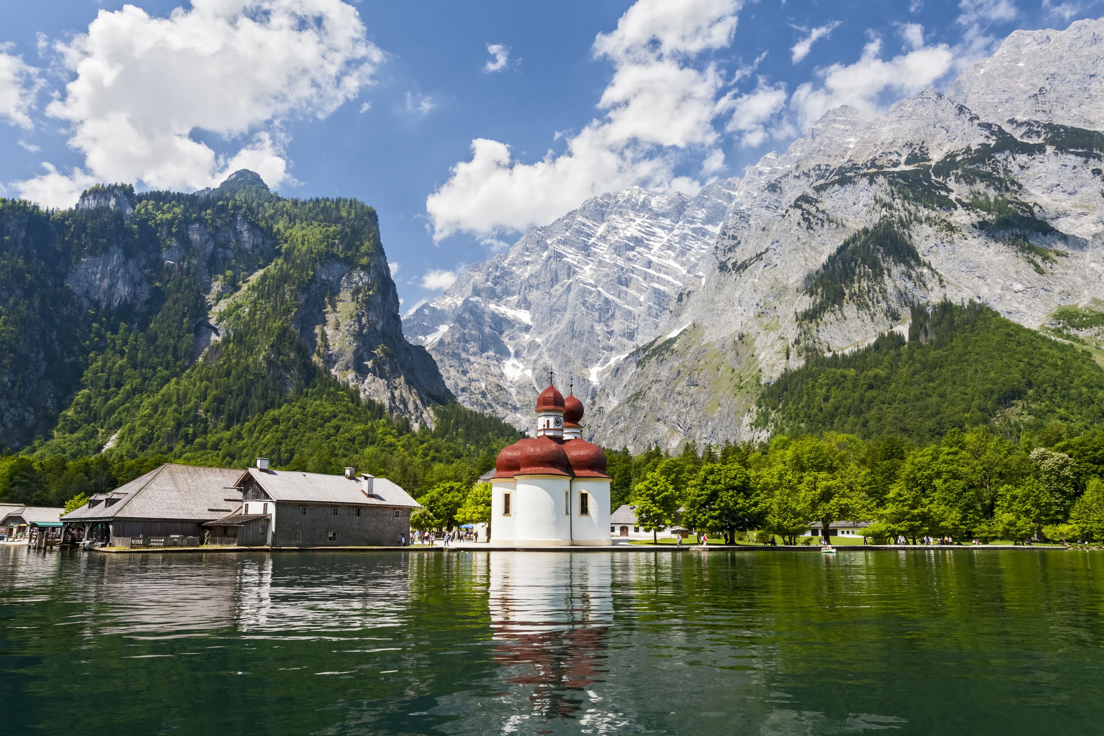 A serene lake with a church featuring a red dome, surrounded by lush greenery and mountains in the background.