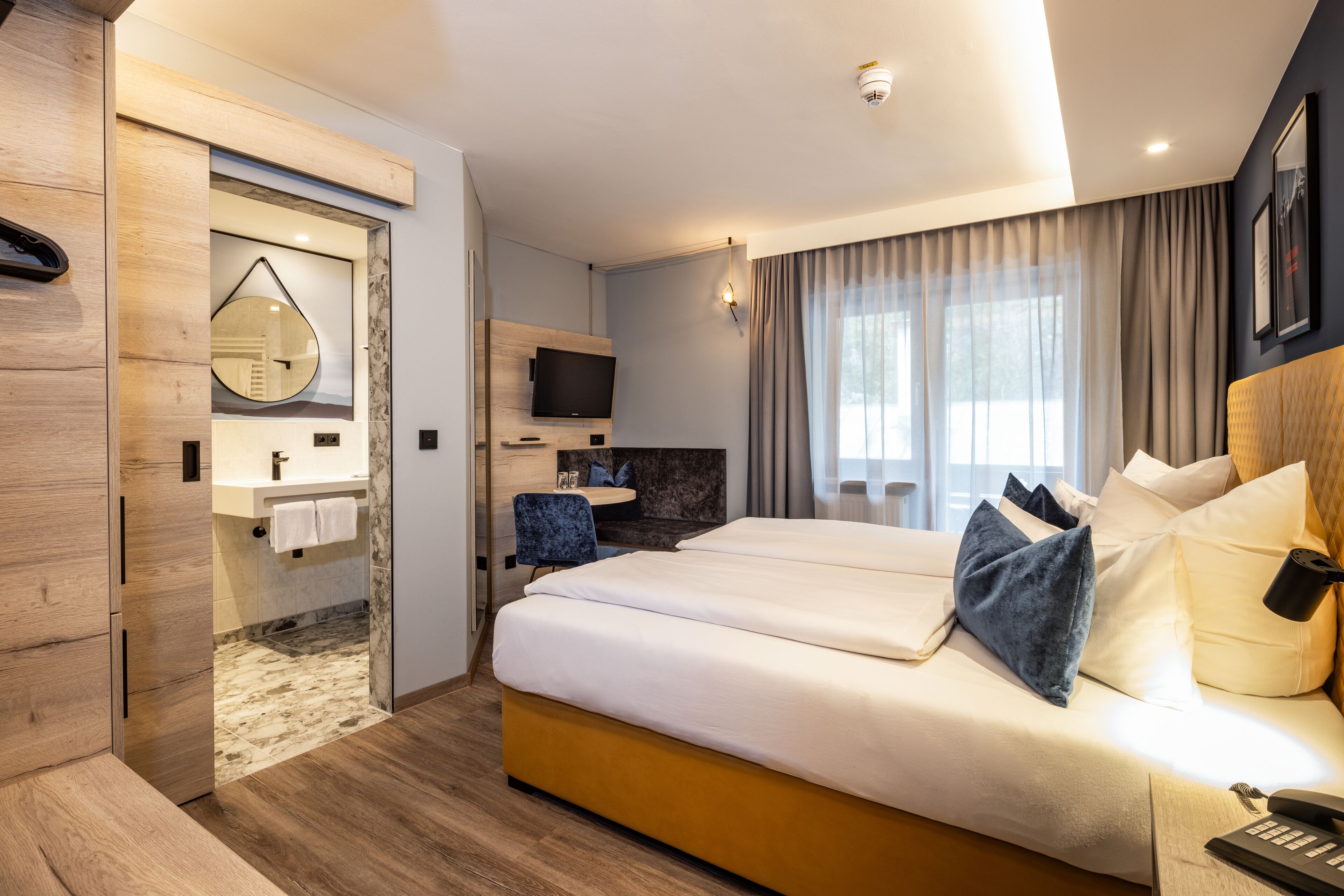 A hotel room with a large bed, a TV, a bathroom with a mirror, and a wooden floor.