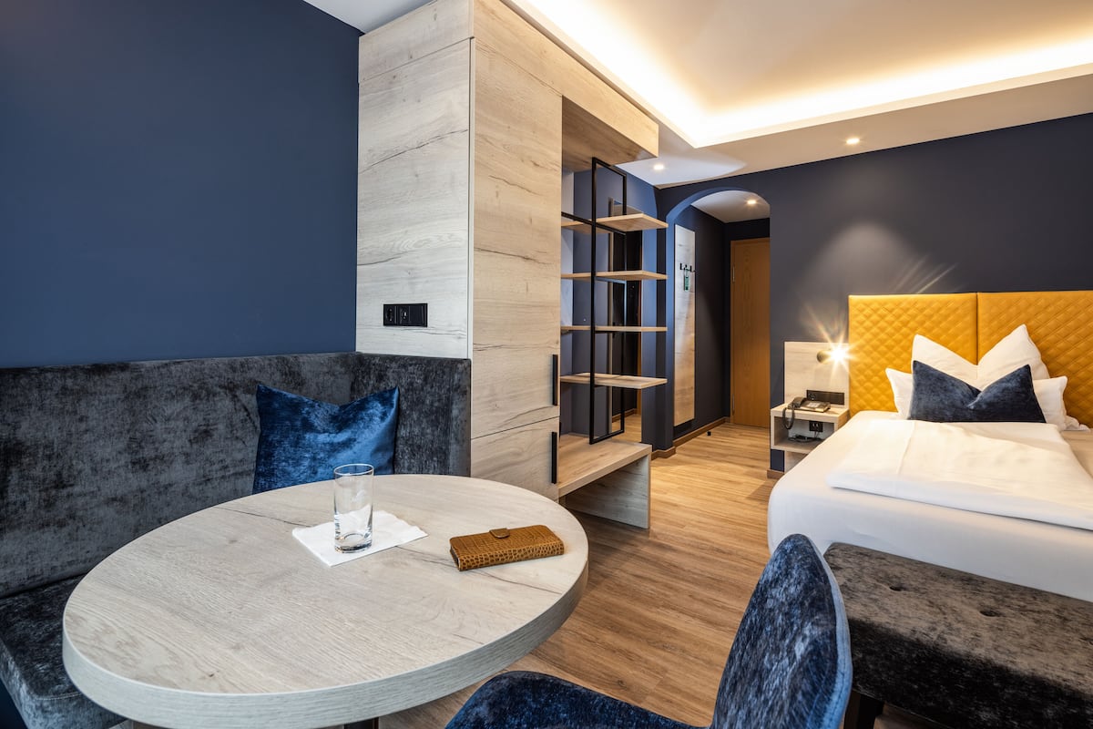 A modern hotel room with a round dining table, a sofa, a bed with a quilted headboard, and a nightstand with a lamp.