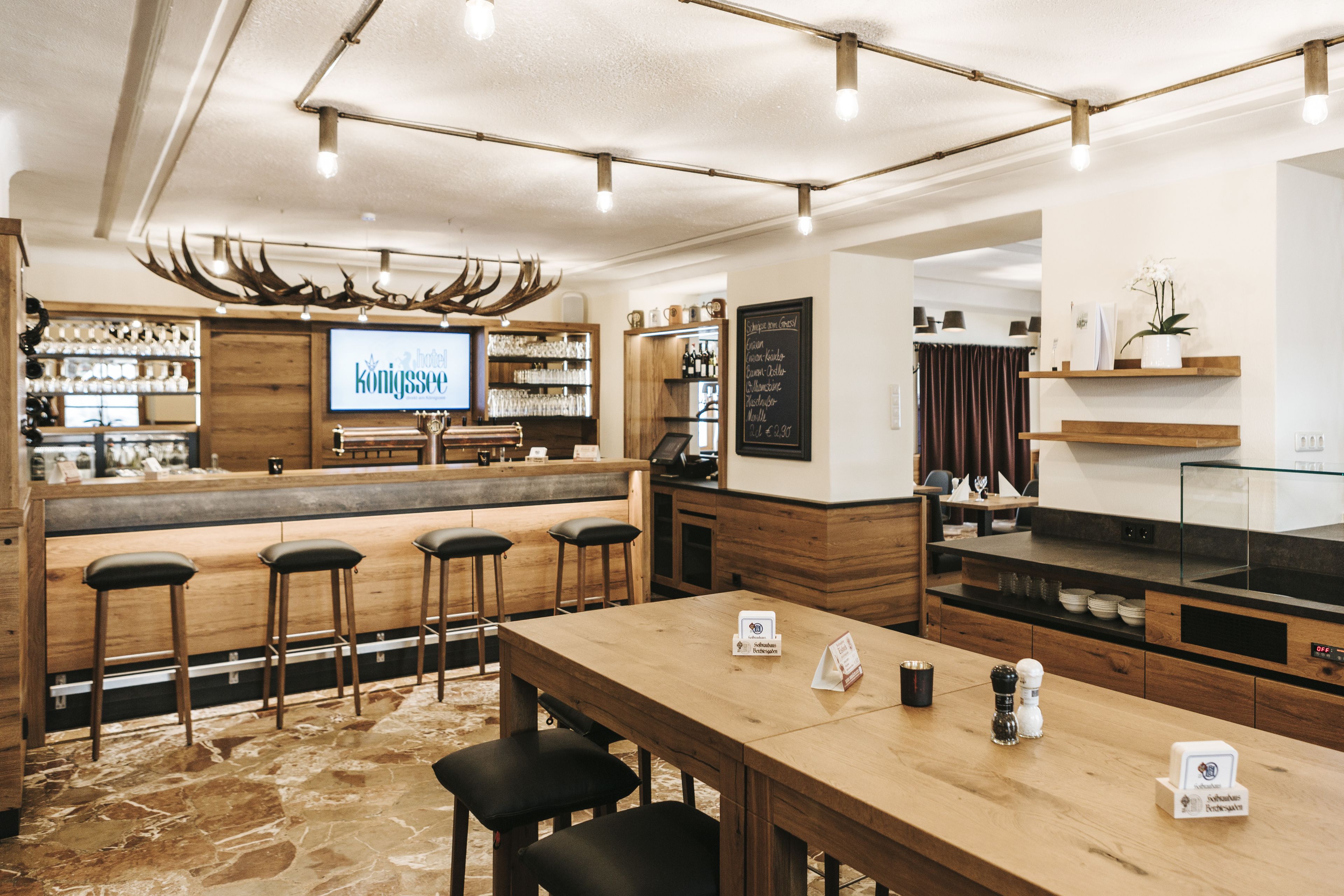 A bar area with wooden stools, a long table, and a television screen displaying a menu.