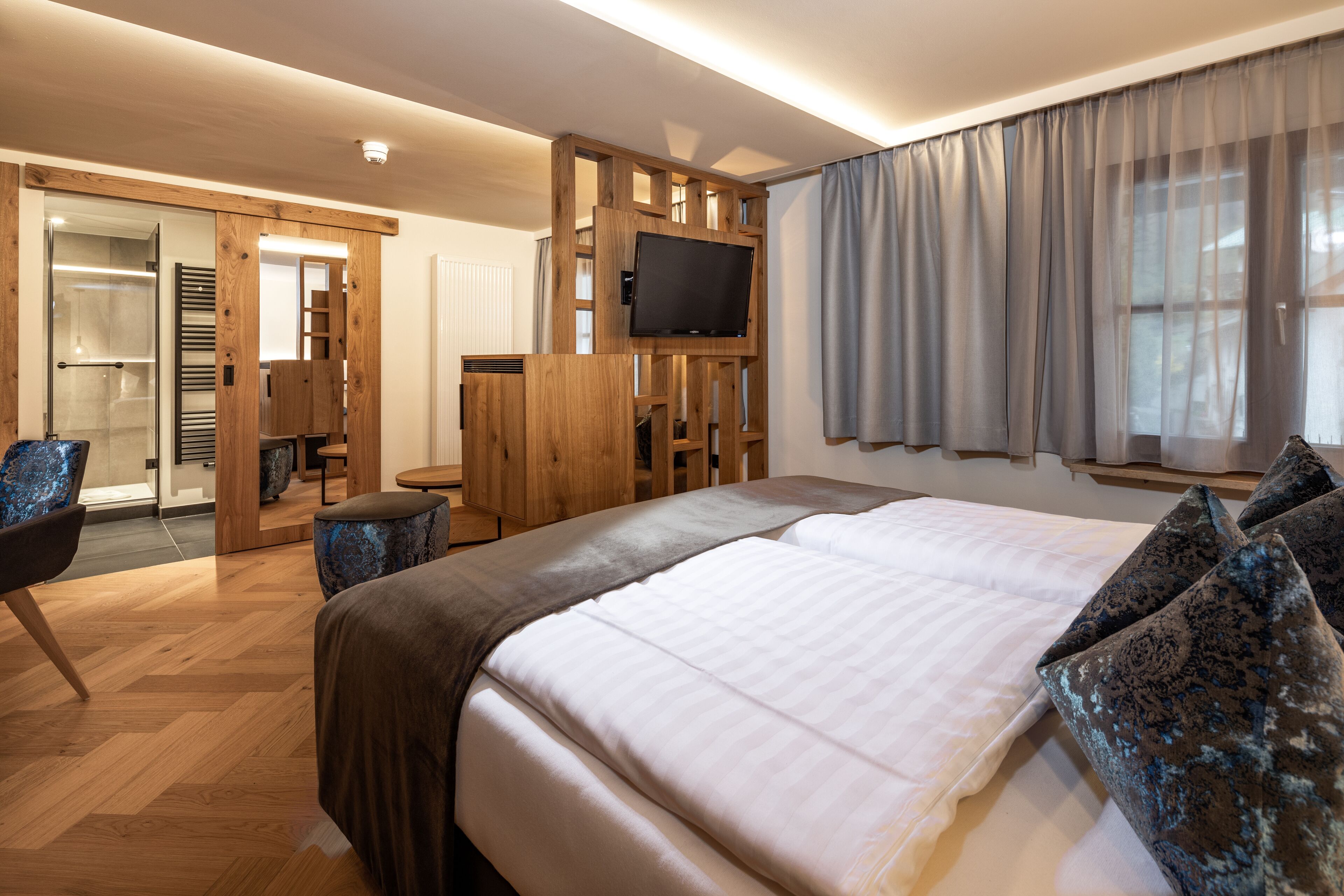 Una habitación de hotel moderna con una cama grande, un televisor montado en un panel de madera, un armario y un espacio de descanso con una silla y una mesa.