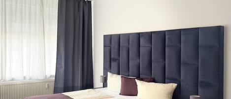 A modern bedroom with a large bed, a nightstand, a bedside lamp, and a window with curtains.