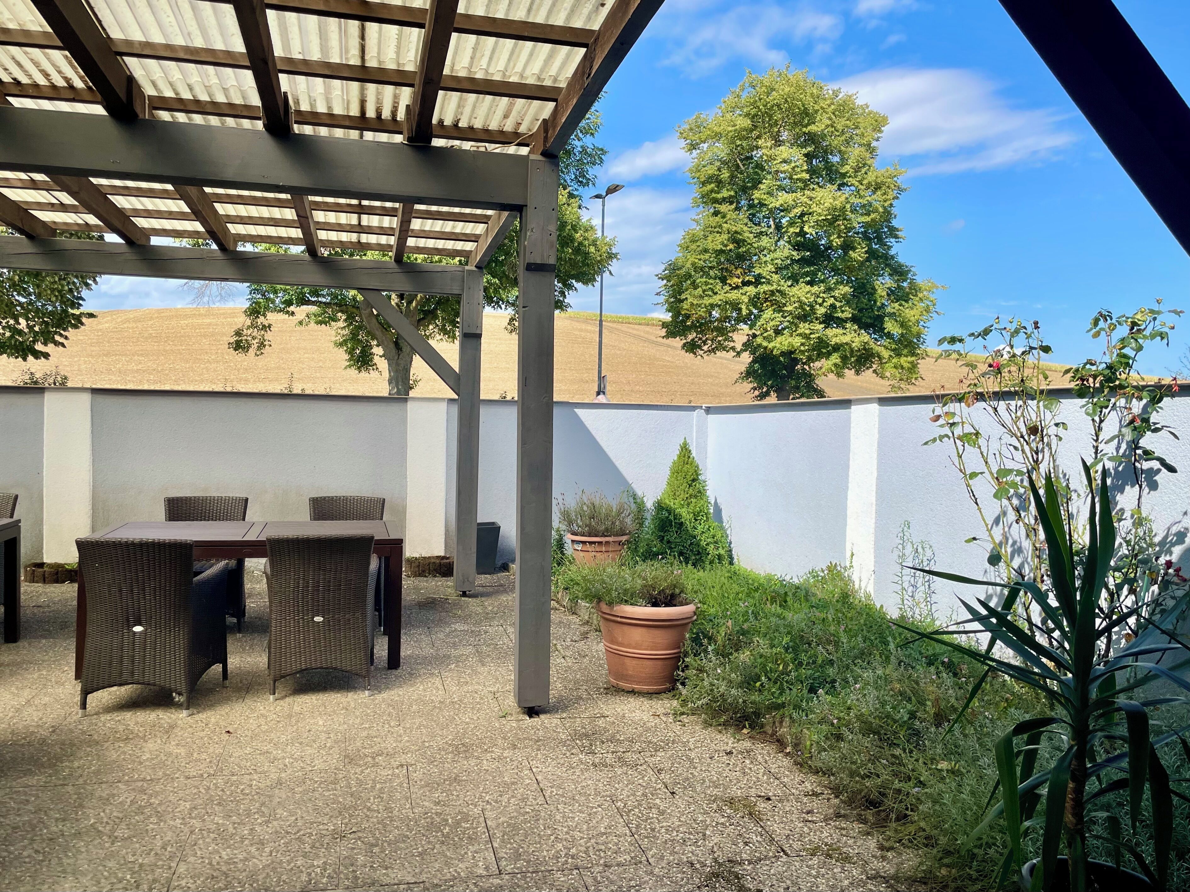 Un patio cubierto con una mesa y sillas, rodeado de un muro blanco y follaje.