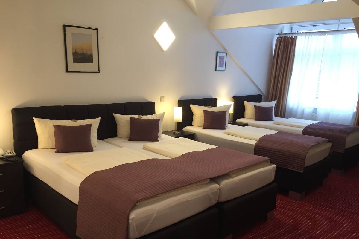 Une chambre d’hôtel avec trois lits, chacun doté d’une tête de lit noire et de draps blancs. Un tableau encadré est accroché au mur, il y a une lampe de chevet et une fenêtre avec des rideaux.