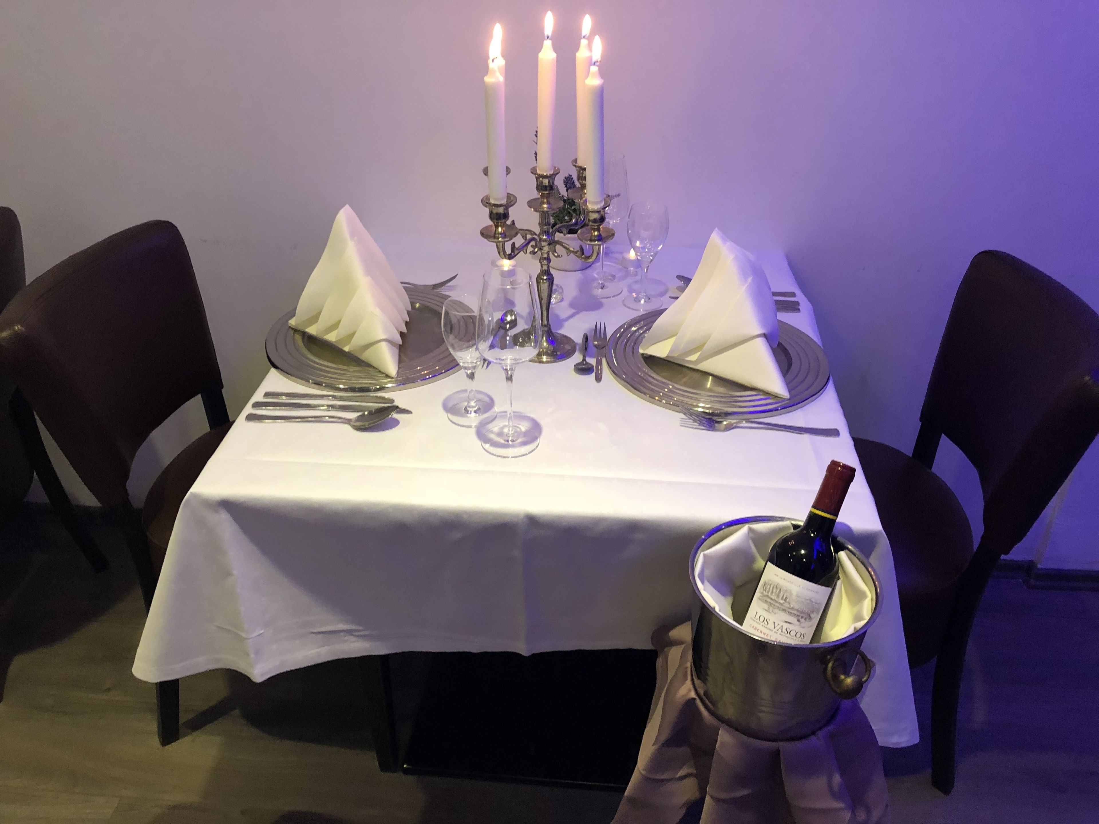 A dining table set with white tablecloth, silverware, wine glasses, and a bottle of wine in a bucket.