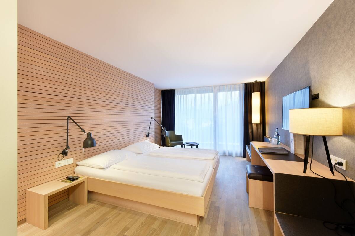 A modern hotel room with a wooden wall, a bed, a desk, a chair, and a TV.