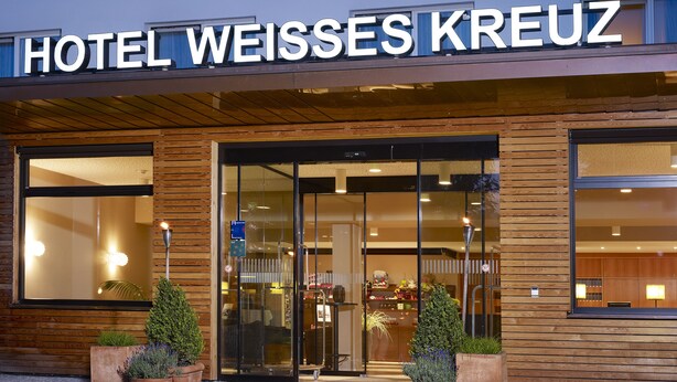 The exterior of Hotel Weisses Kreuz with a modern wooden facade, large glass entrance, and potted plants in front.