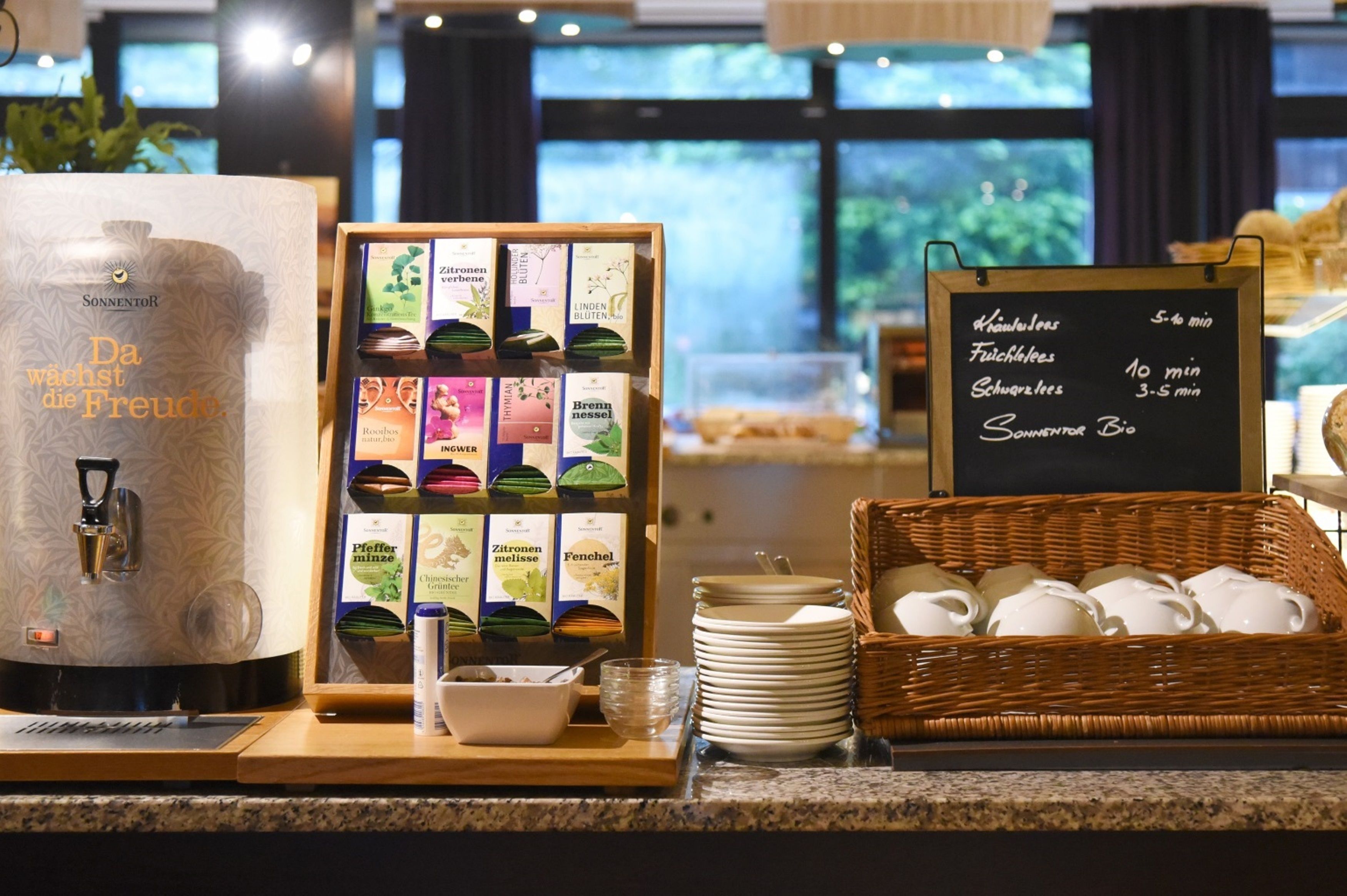 Una estación de café con dispensador de agua, sobres de té y café, y un panel con el menú.