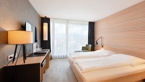 A modern hotel room with a wooden bed, a desk with a lamp, a chair, and a large window with sheer curtains.