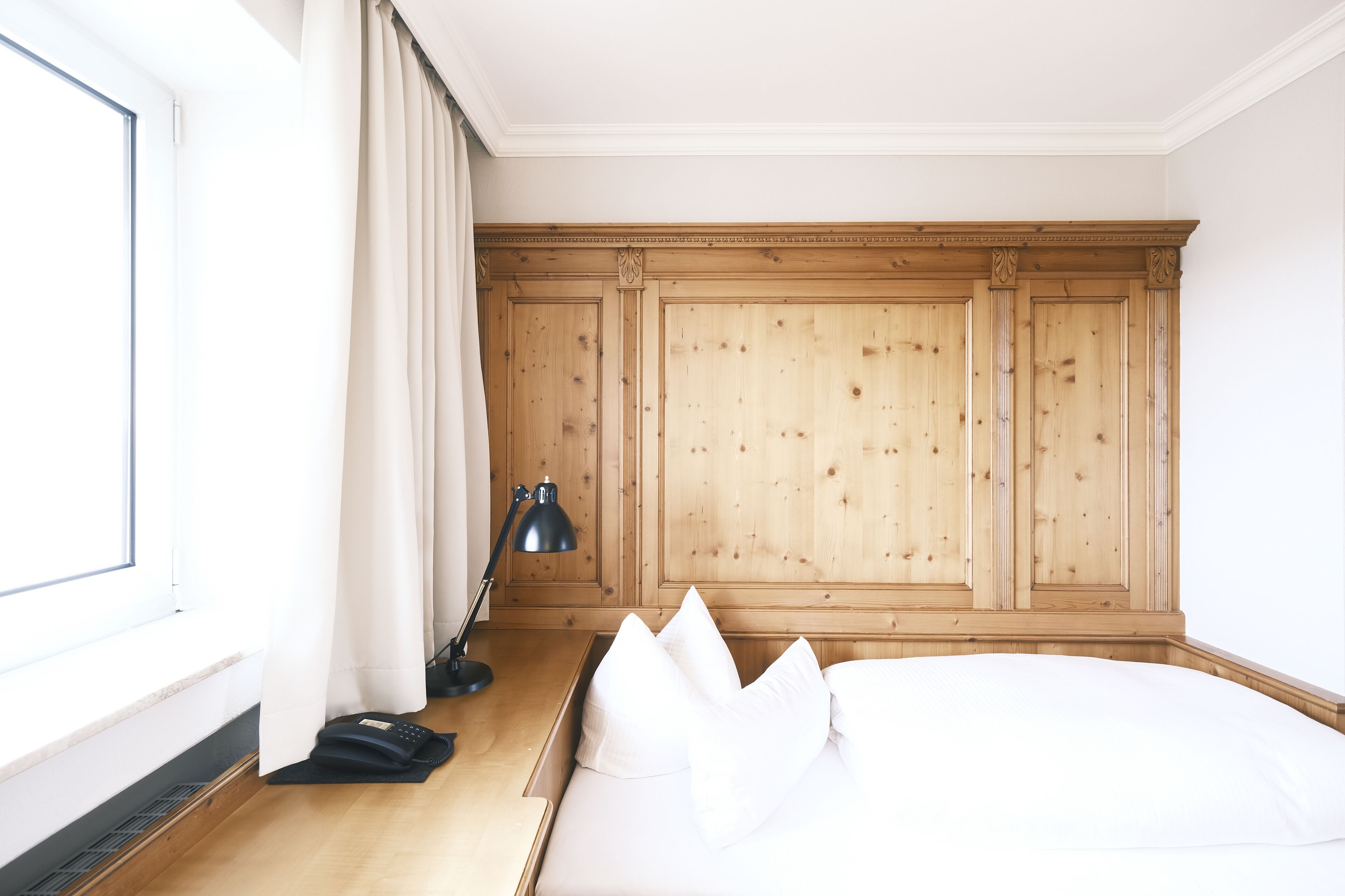 Un dormitorio con cabecera de madera, cama con ropa de cama blanca, mesita de noche con lámpara y ventana con cortinas.