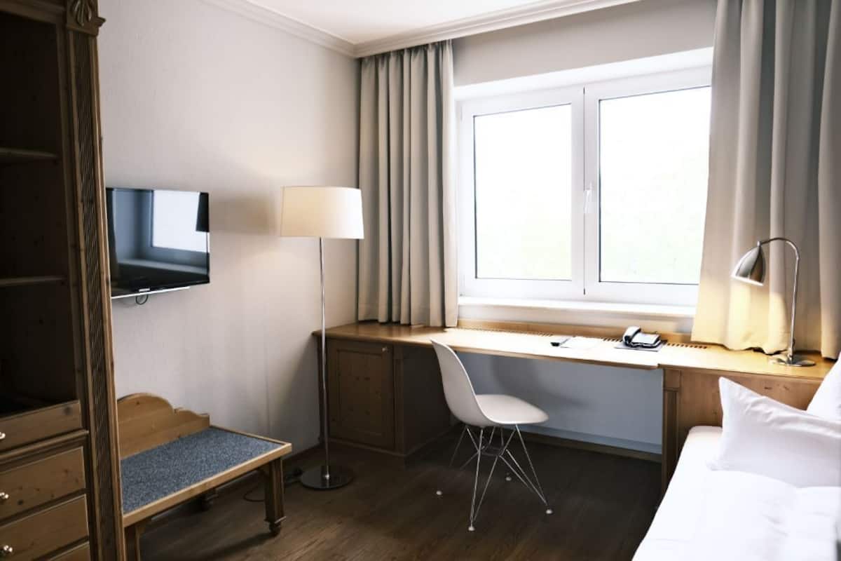 A hotel room with a wooden desk, a white chair, a TV mounted on the wall, a bedside lamp, and a window with curtains.
