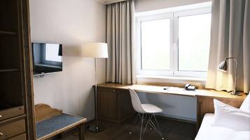 A hotel room with a wooden desk, a white chair, a TV mounted on the wall, a bedside lamp, and a window with curtains.