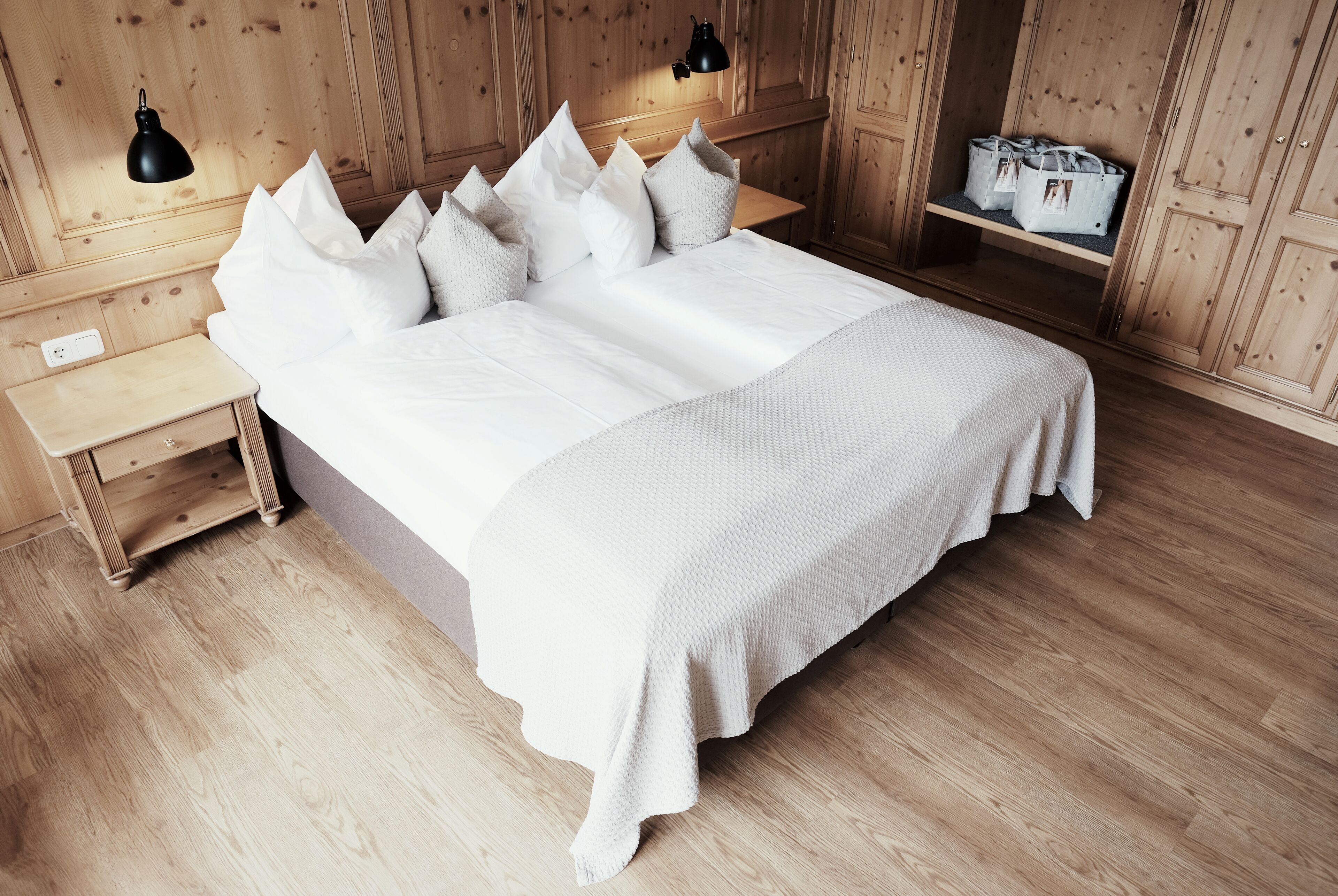 A neatly made bed with white linens and a matching throw blanket, wooden headboard, bedside lamps, and a wooden nightstand with a drawer.