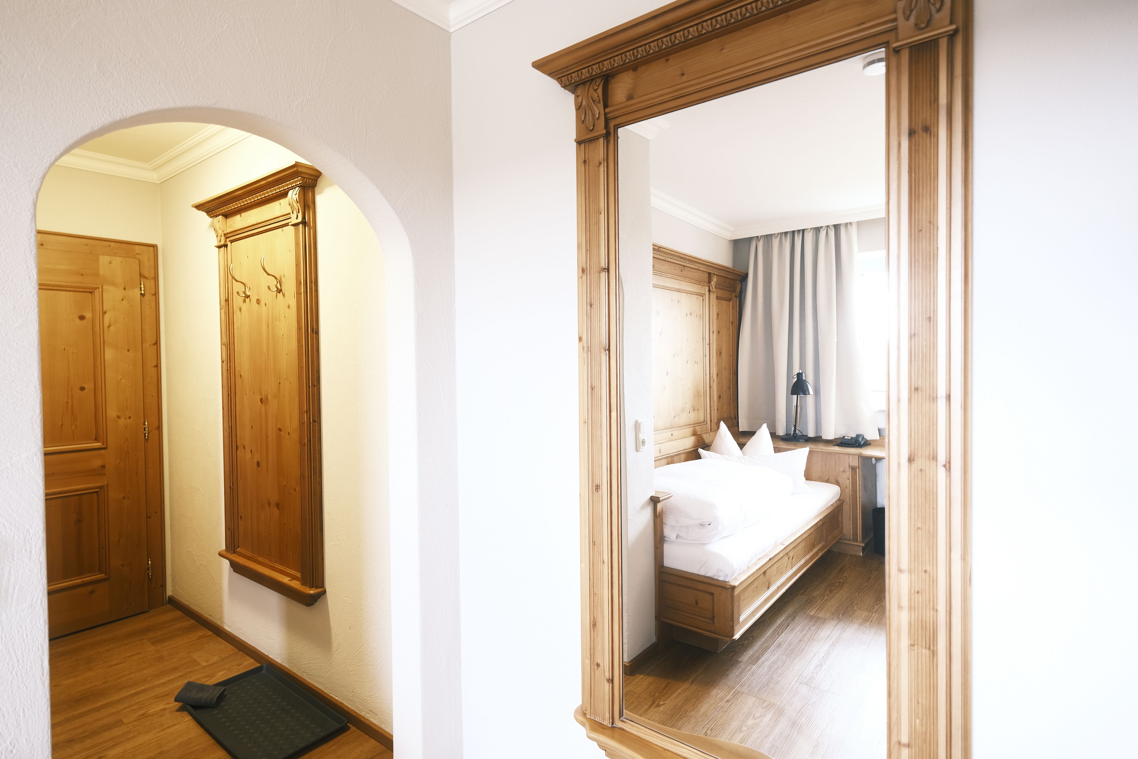 A hotel room with a wooden wardrobe, a bed with white linens, and a large mirror reflecting the room.
