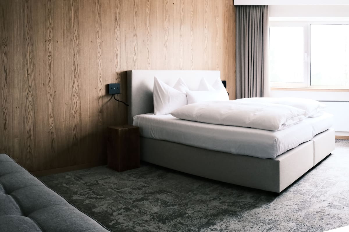 A modern bedroom with a wooden wall, a grey sofa, a bed with white bedding, and a window with curtains.