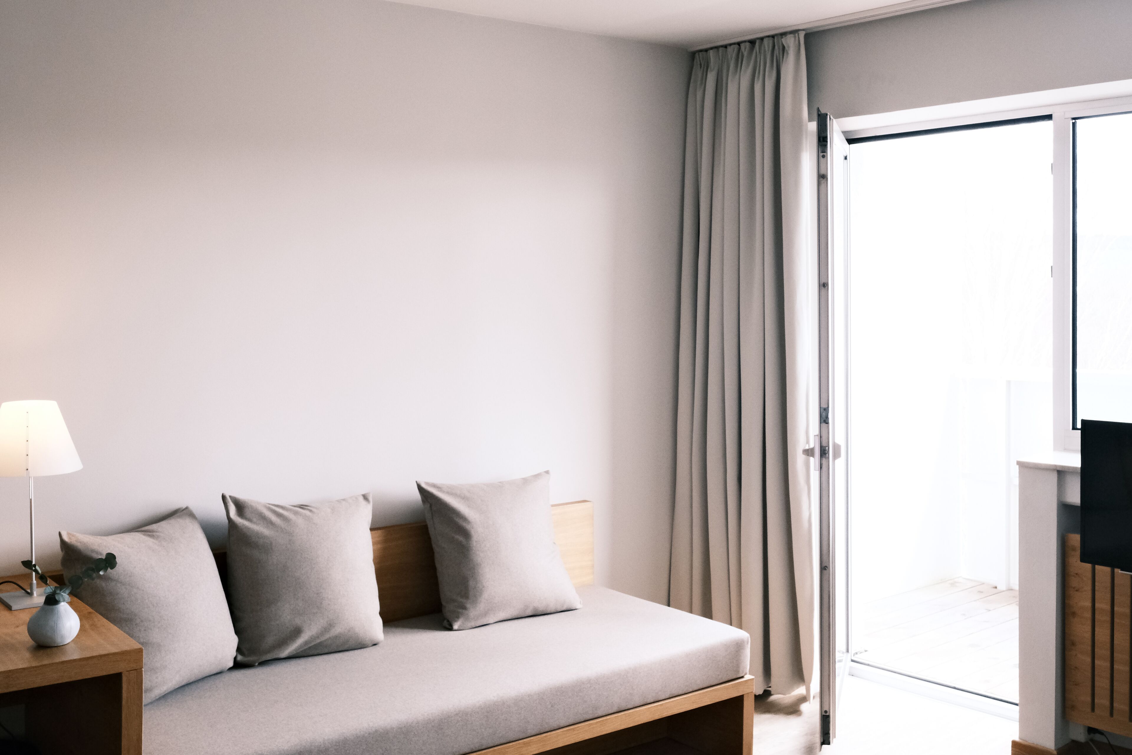 Una habitación de hotel moderna con sofá cama, una mesita de noche con lámpara y televisor sobre un mueble de madera.
