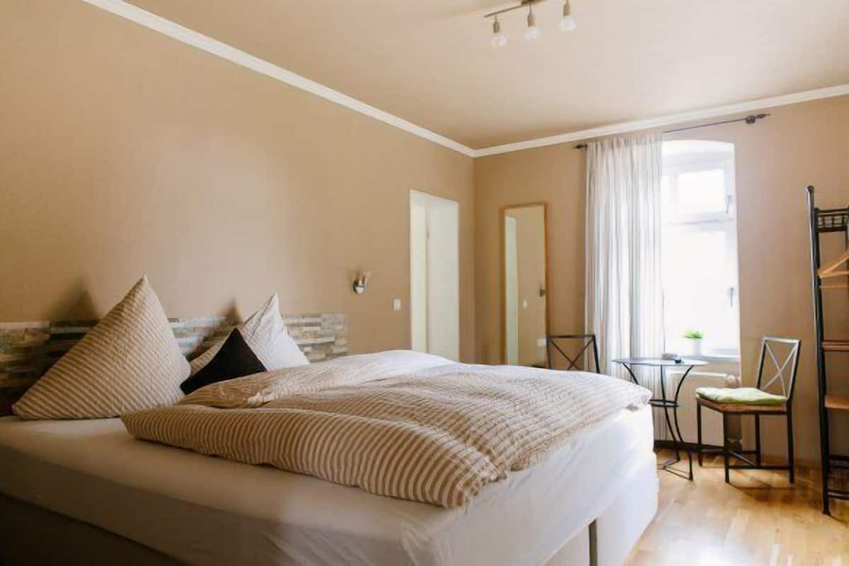 Un dormitorio con cama, una silla, una mesita y una ventana con cortinas.