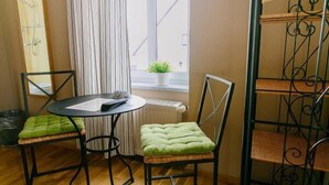 Una habitación pequeña con una mesa redonda, dos sillas con cojines verdes, una ventana con cortinas, un radiador y una escalera de metal.
