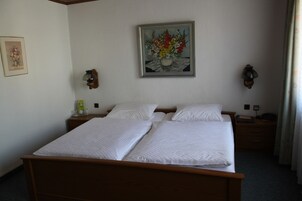 A double bed with white linens, a nightstand with a lamp, and a painting of flowers on the wall.