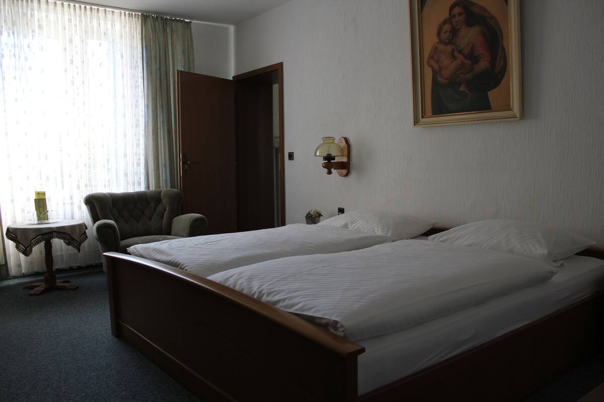 A hotel room with a double bed, a chair, a small table, and a painting on the wall.
