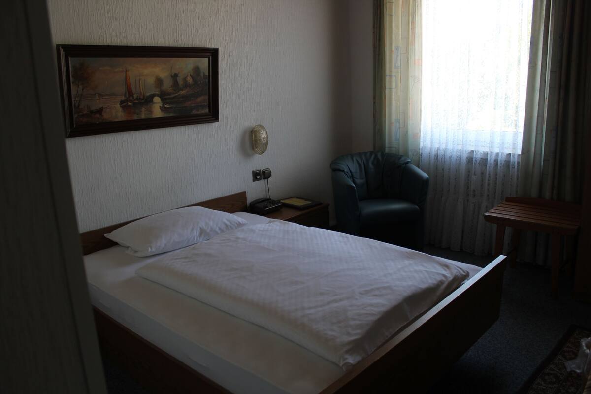 A hotel room with a bed, a chair, a painting on the wall, and a window with curtains.