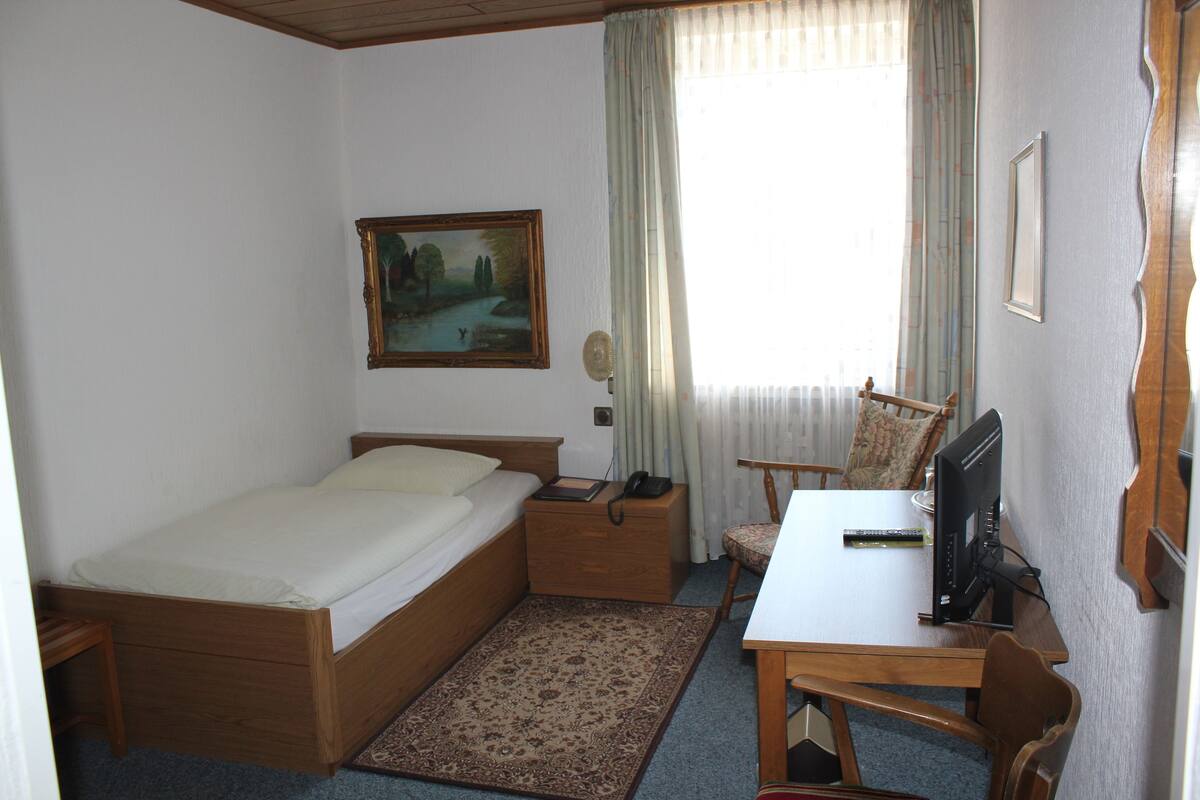 A bedroom with a single bed, a desk with a computer, a chair, a nightstand, a painting, and a window with curtains.