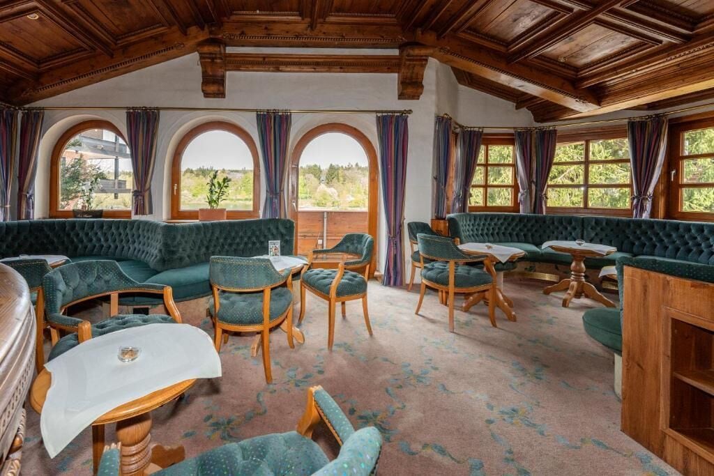 A room with a high ceiling, wooden beams, green upholstered seating, and large windows with curtains.