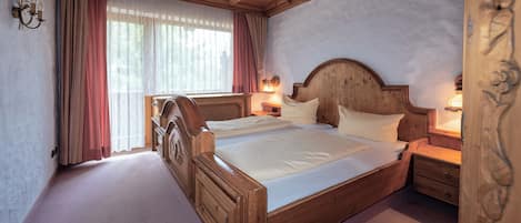 Ein Doppelbett mit hölzernem Kopfteil und Fußteil, zwei Nachttische mit Lampen, ein großes Fenster mit leichtem Vorhang und eine Holzbalkendecke.